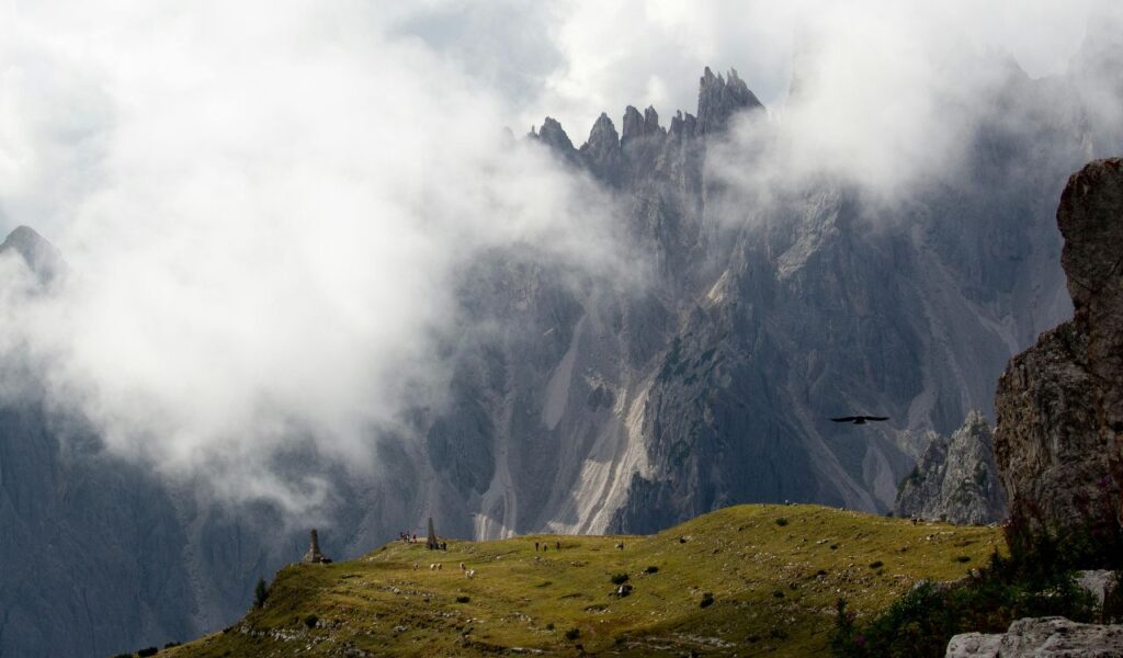 **Alt text:**
Grassy alpine plateau in Val di Funes with scattered hikers and a stone monument, set against dramatic, jagged Odle peaks partly shrouded in clouds, with a bird soaring in the foreground.