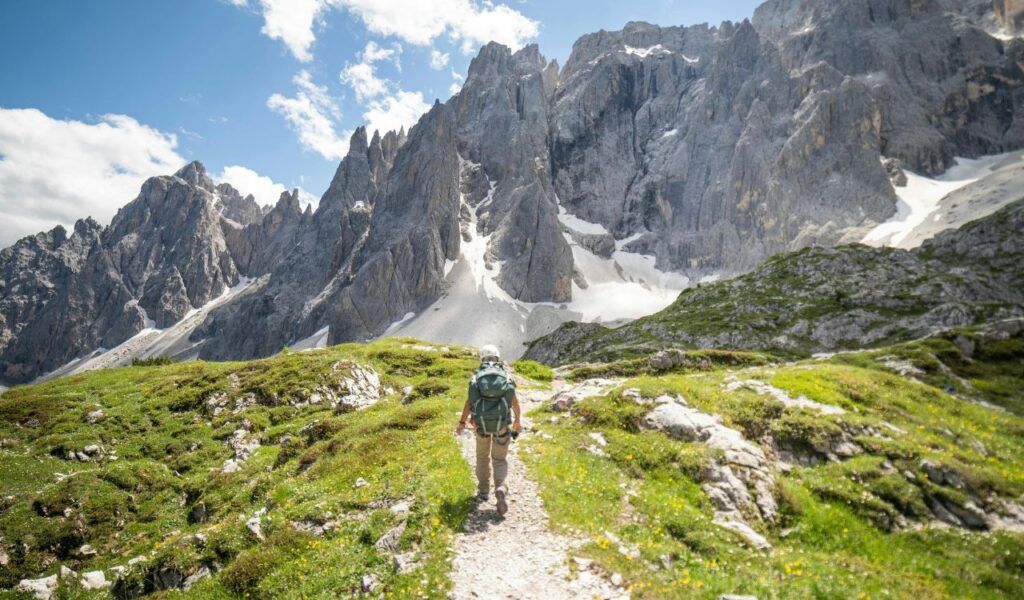 **Alt text:**
A hiker with a backpack walks along a narrow mountain trail surrounded by lush green grass and wildflowers, heading toward the jagged, snow-patched peaks of the Dolomites under a bright blue sky.