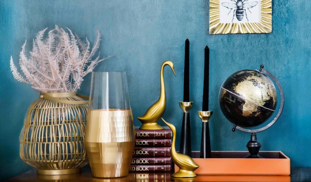 Decorative shelf with gold vases, black candles, globe, books, and metallic bird figurines against a teal wall.