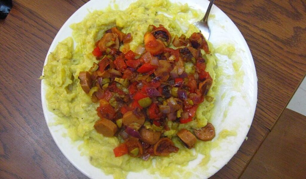 Plate of mashed potatoes topped with grilled vegetables and sausage slices, served on a wooden table.