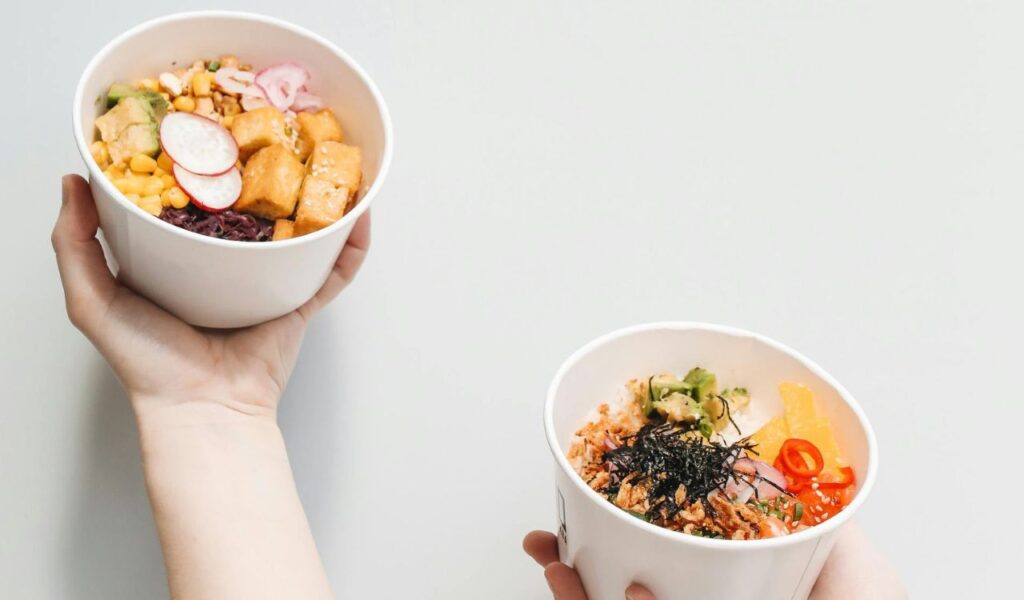 Two hands hold bowls of colorful fresh food, topped with vegetables, tofu, seaweed, and garnishes, against a plain white background.