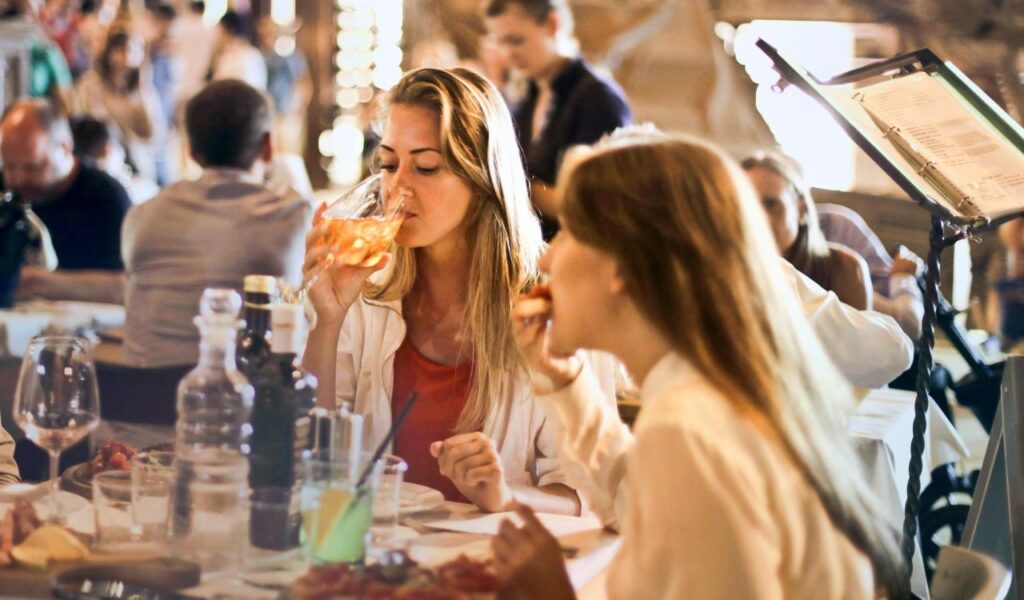 Two women enjoy drinks and food at a lively, warmly lit restaurant, surrounded by other diners and a casual, social atmosphere.