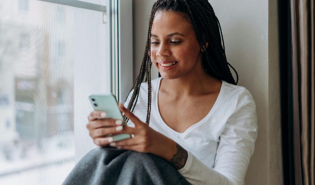 A woman sitting by a window, smiling while using her smartphone, possibly adjusting phone settings or browsing.