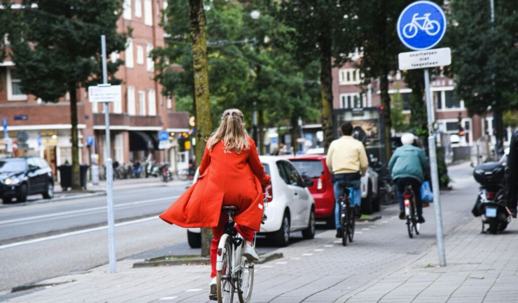 Woman in a bright red coat riding a bicycle on a designated bike path in an urban area, surrounded by other cyclists and city traffic.