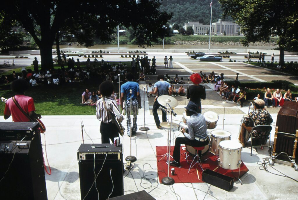 Band performing outdoors in the 1970s with vintage instruments and clothing, facing a seated crowd near a river and government building.