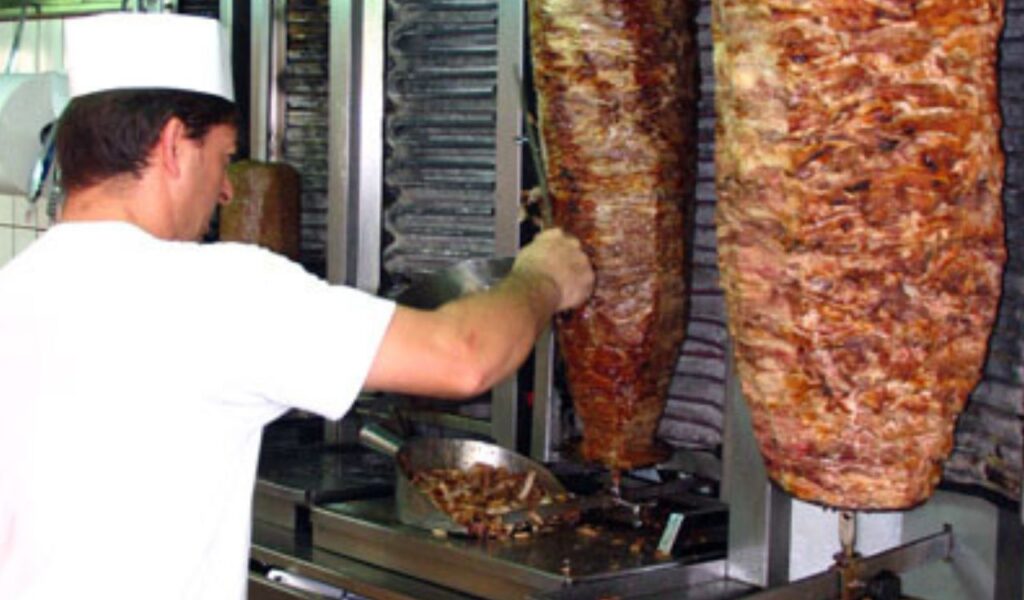 Man slicing meat from a vertical gyro rotisserie in a traditional Greek eatery, preparing fresh gyro servings.