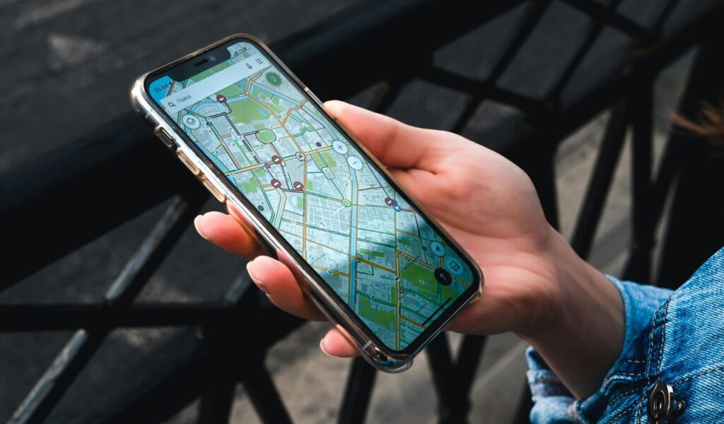 A person holding a smartphone displaying a detailed offline map app, viewed in daylight with a street railing in the background.