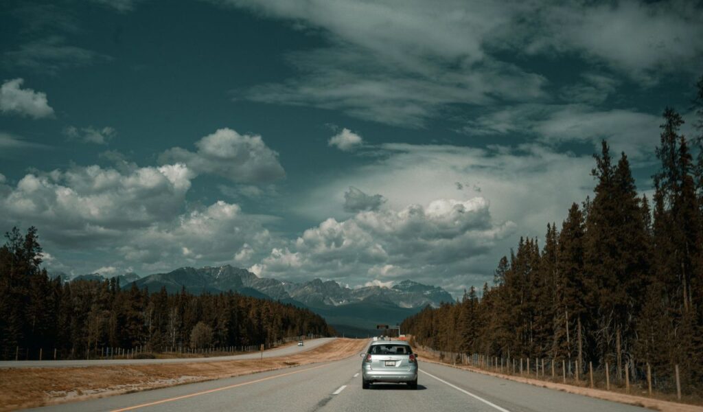 A silver car drives along a scenic highway surrounded by pine trees and distant mountains under a dramatic sky filled with clouds, evoking the feel of a mountain road trip.