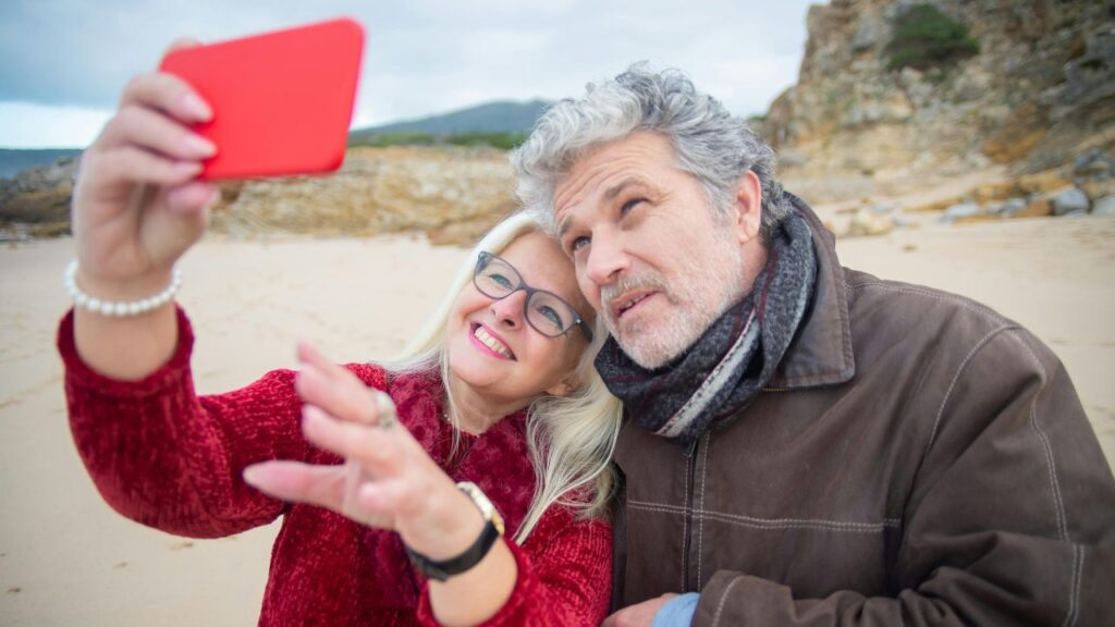 Smiling older couple taking a selfie at the beach, dressed warmly with rocky cliffs and sand in the background.