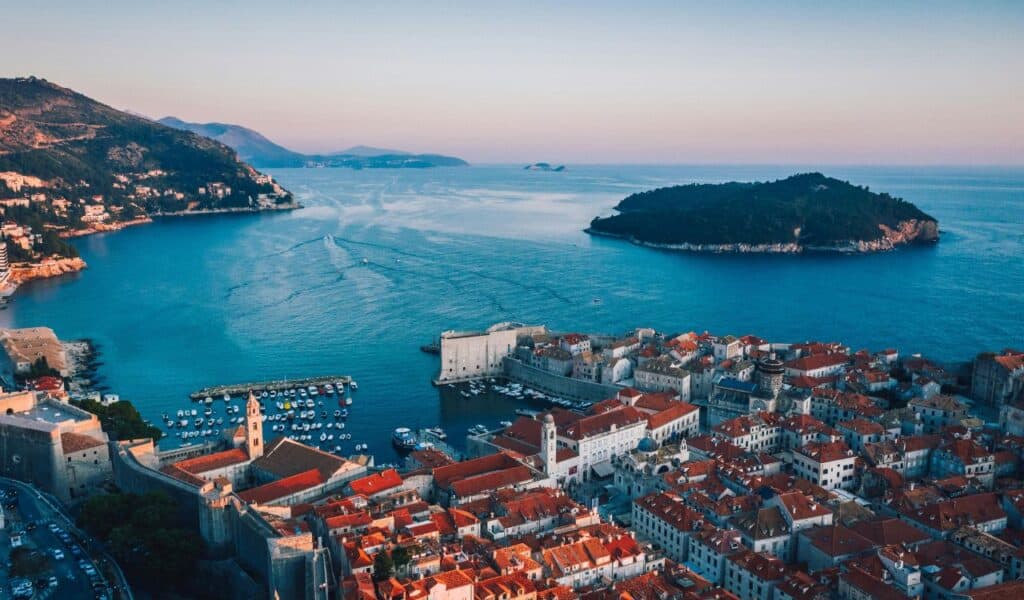 Aerial view of Dubrovnik’s old town with its terracotta rooftops, historic city walls, and harbor, overlooking the Adriatic Sea and a lush green island at sunset.
