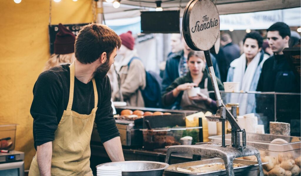 A vendor wearing a yellow apron prepares food at a busy outdoor market stall called "The Frenchie" while customers wait in line to be served.