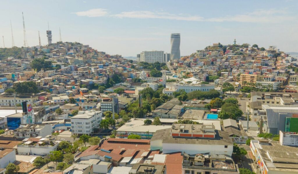 Aerial view of Guayaquil, Ecuador, showcasing colorful hillside houses, modern high-rise buildings, and urban streets under a bright blue sky.