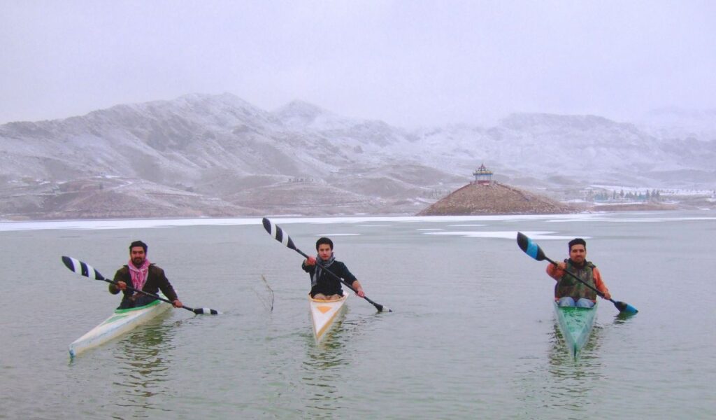 Three men kayaking on a cold lake with snowy mountains and a pagoda-style hut in the background.