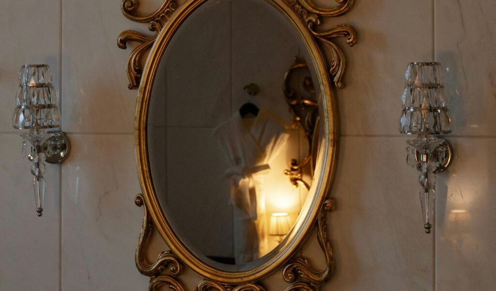 White hotel bathrobe reflected in an ornate oval mirror, flanked by two crystal wall sconces in a tiled bathroom.