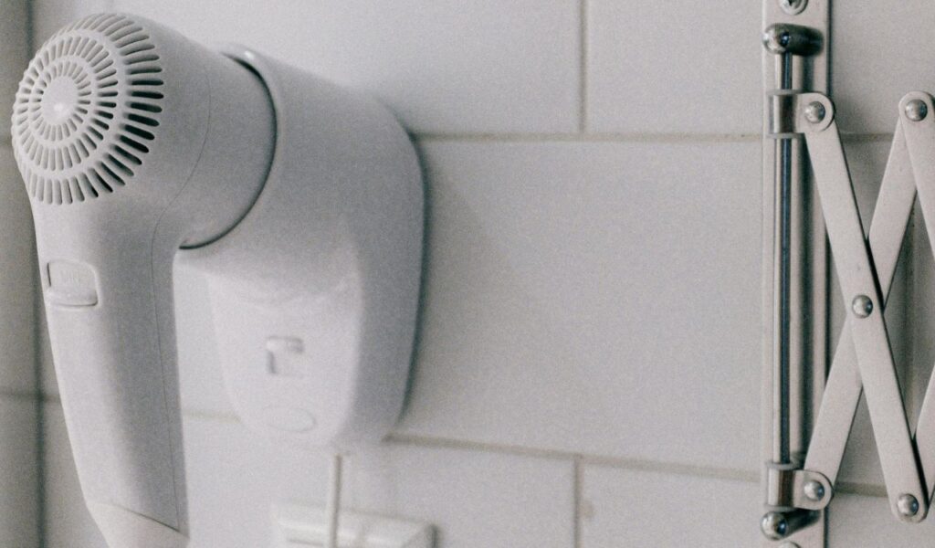 Wall-mounted white hair dryer in a tiled hotel bathroom, positioned next to a metal accordion-style mirror.