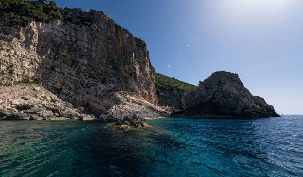  Rugged coastal cliffs rise above clear blue waters under a bright sky, with rocky outcrops and lush greenery in the background.
