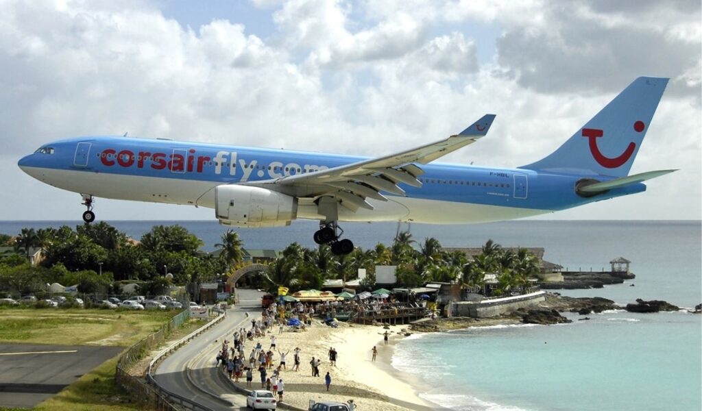 Airplane flying low over a beach, with tourists walking on the sand below, near a tropical destination.