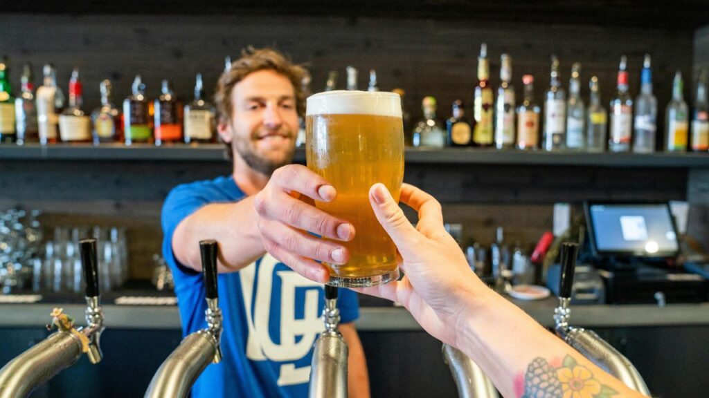 Bartender smiling while handing a fresh pint of beer across the bar to a customer with a floral tattoo on their arm.