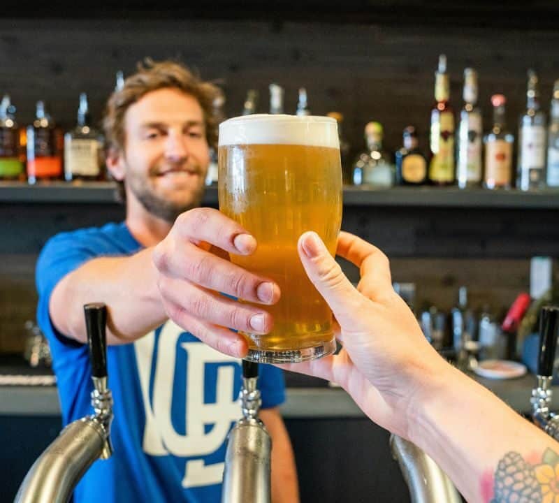 Bartender smiling while handing a fresh pint of beer across the bar to a customer with a floral tattoo on their arm.
