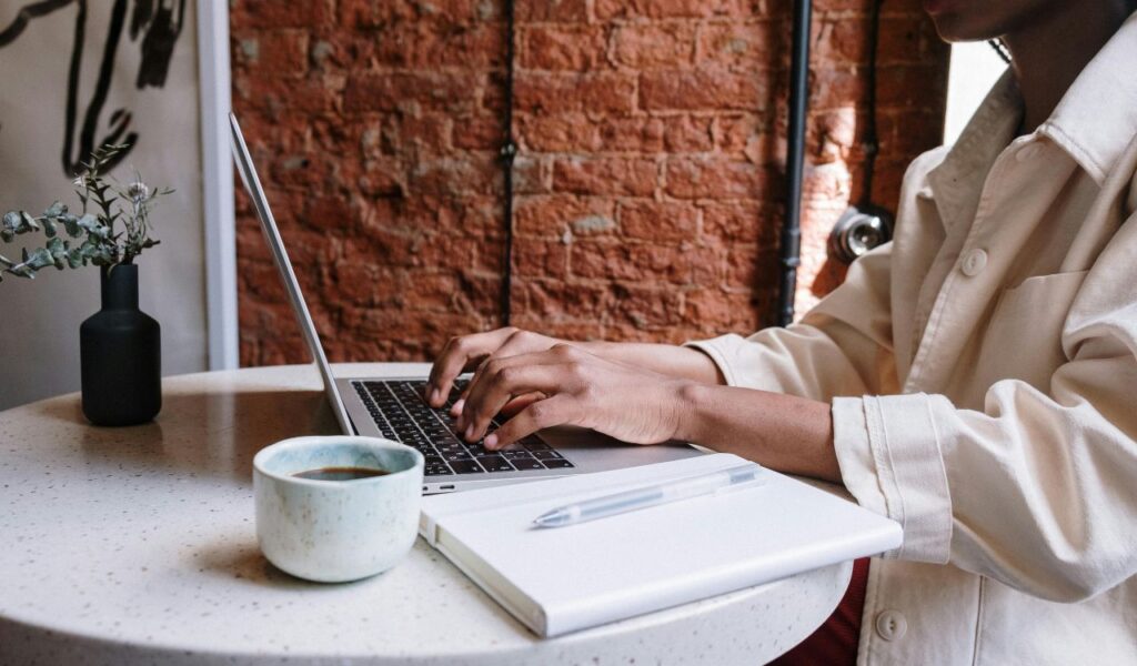 Person typing on a laptop at a café table with a coffee mug and notebook nearby, suggesting public Wi-Fi use and potential exposure to cyber threats like fake networks.