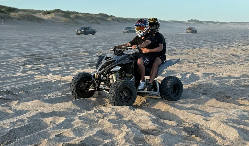 Two people wearing helmets ride an ATV across a sandy beach, with off-road vehicles in the background and sand dunes lining the horizon.