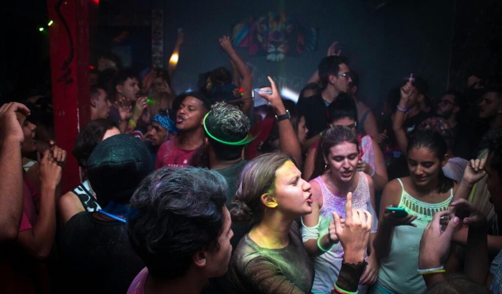 Young people dance and sing in a crowded, energetic nightclub with colorful lights and glow accessories, creating a lively party atmosphere.