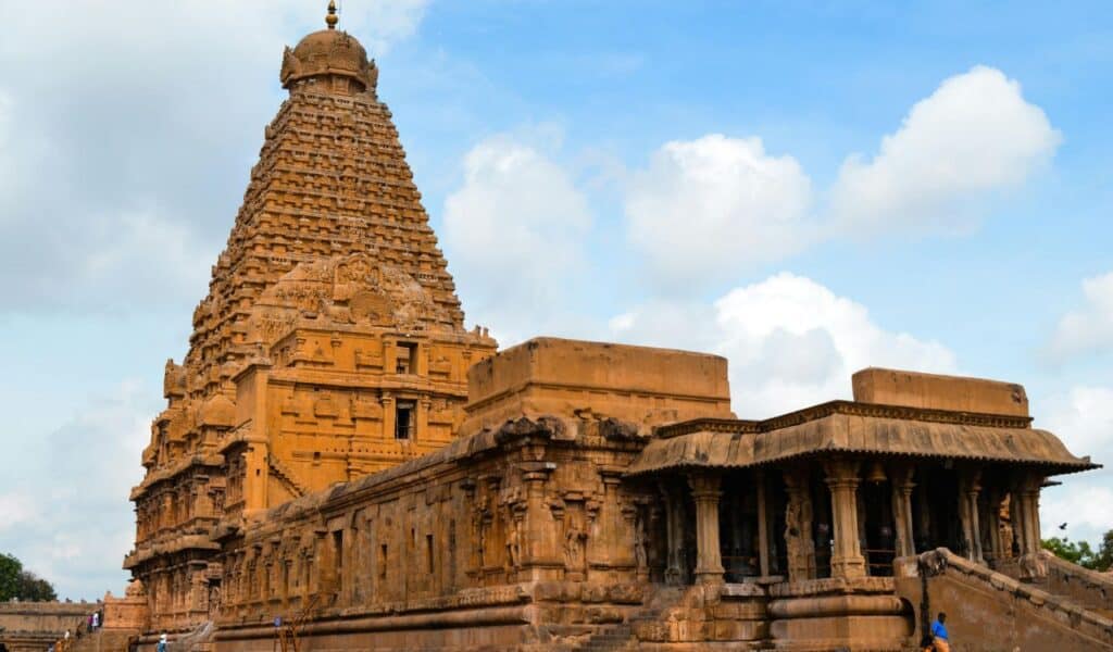 Majestic stone temple with a towering, intricately carved vimana and pillared mandapa, set against a bright blue sky with scattered clouds in Tamil Nadu, India.