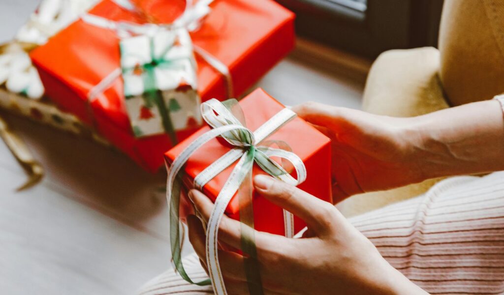 A person holding a small red gift box with a green and white ribbon, with more wrapped presents in the background, suggesting the theme of free gifts or giveaways.
