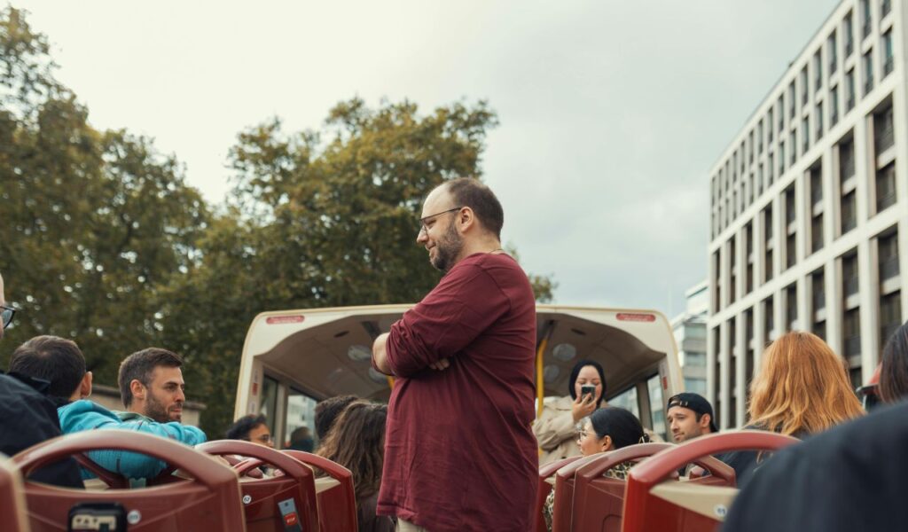A man standing and speaking to passengers on the upper deck of a tour bus, surrounded by seated tourists, suggesting a guided city tour experience.