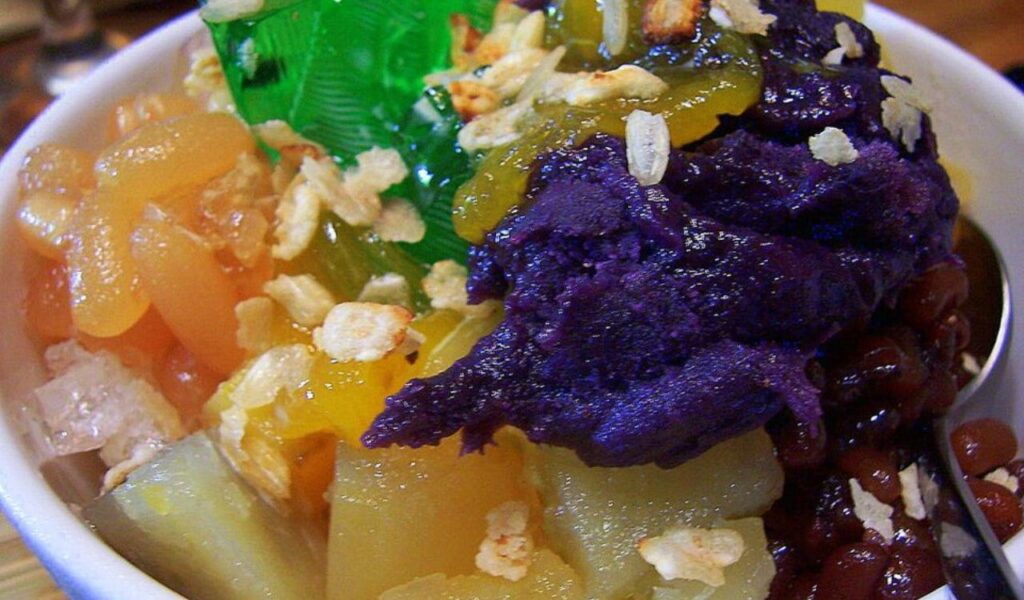 Close-up of Filipino halo-halo dessert with crushed ice, colorful jellies, fruits, beans, and purple yam in a white bowl.