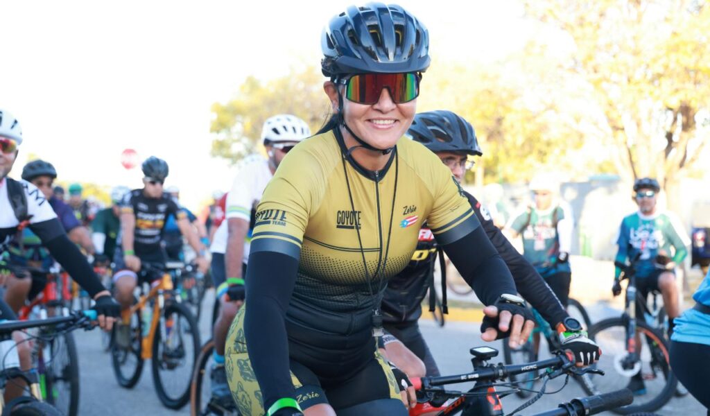 Smiling woman in cycling gear and helmet surrounded by a diverse group of cyclists at the start of a group ride or community event.