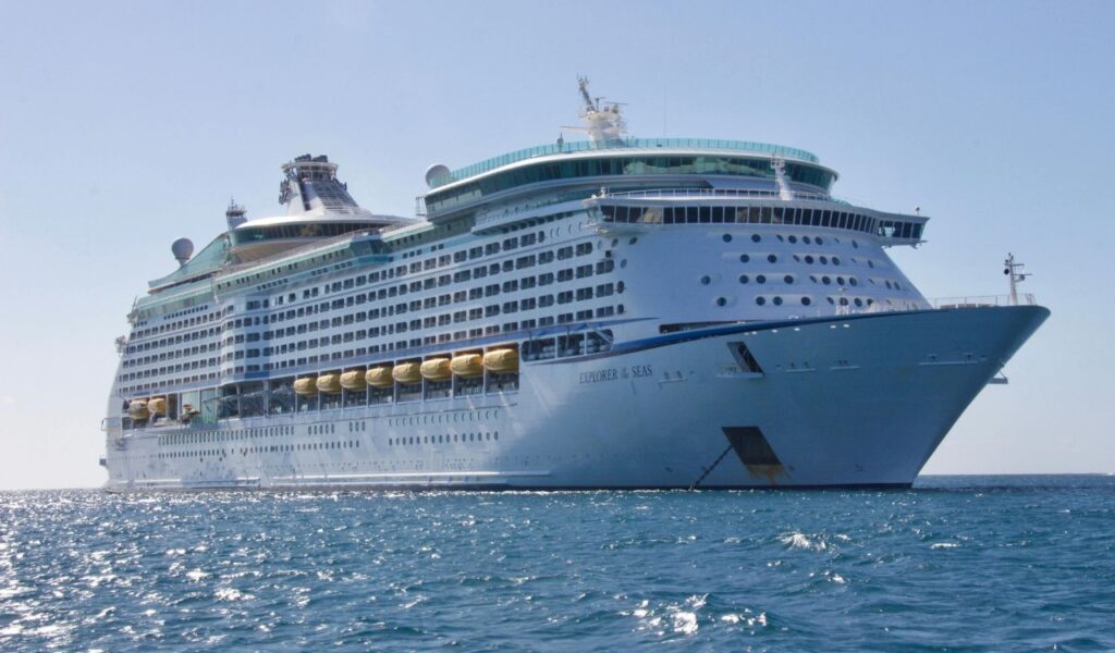 A large cruise ship floats on clear blue ocean water under a sunny sky, showcasing multiple decks lined with windows and balconies.