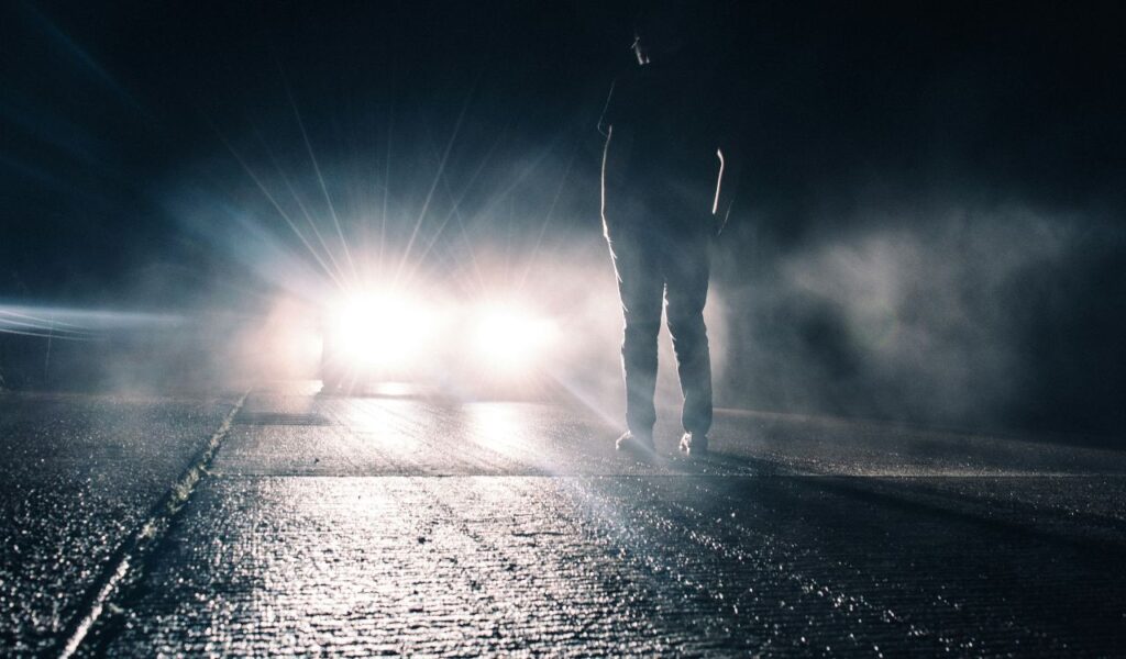A person stands on a dark road facing an approaching car with bright headlights, creating a dramatic silhouette and glare through the foggy night air.