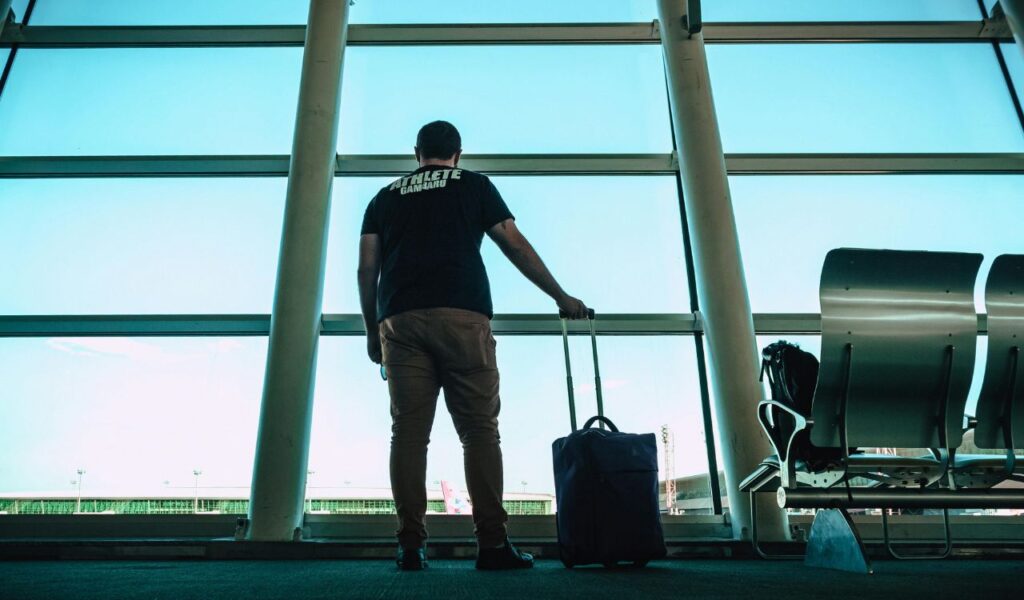 Solo traveler standing with a suitcase at an airport terminal window, gazing out at the runway and airport buildings.
