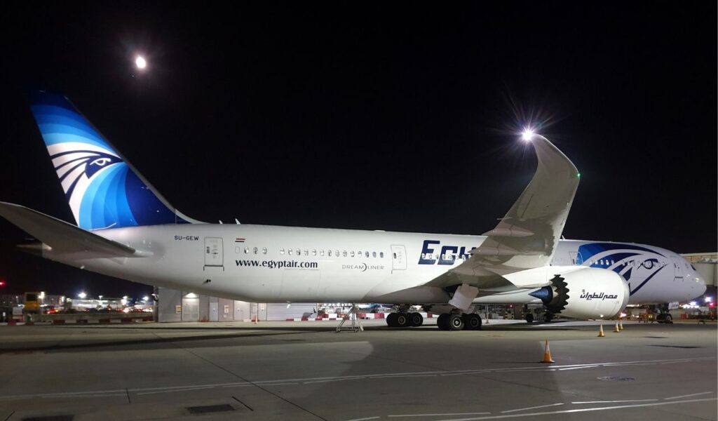 An EgyptAir Dreamliner parked on the tarmac at night under a bright moon, illustrating a red-eye flight option for late-night travel savings.