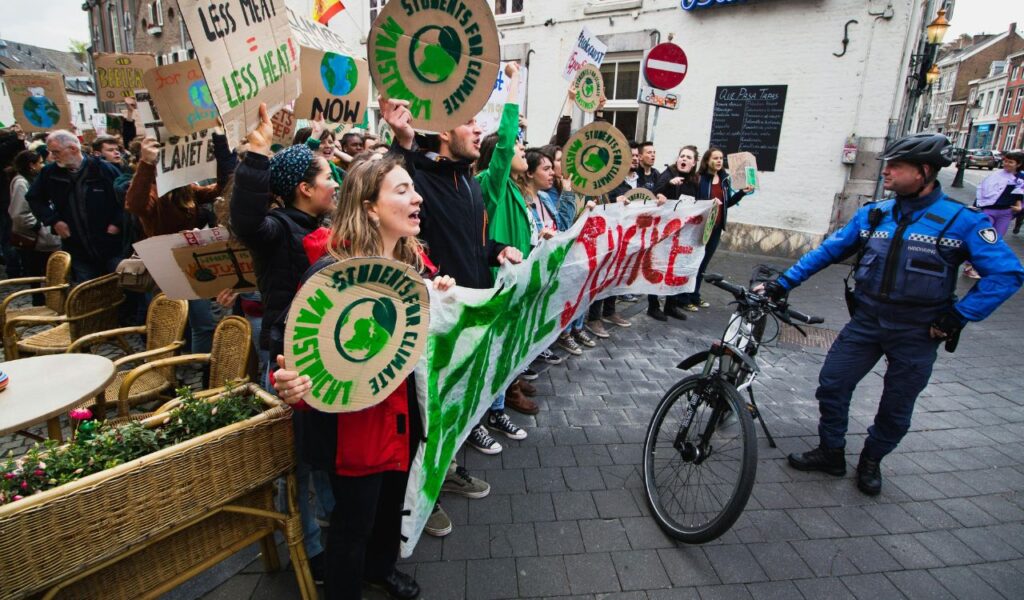 A group of climate activists hold protest signs and a banner reading "Climate Justice" while a police officer with a bicycle stands nearby.