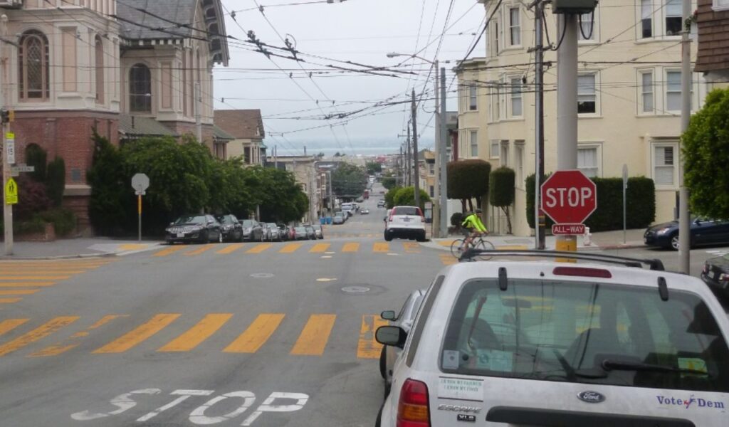 A quiet urban four-way intersection with stop signs on all corners, yellow pedestrian crossings, and parked cars lining the street.
