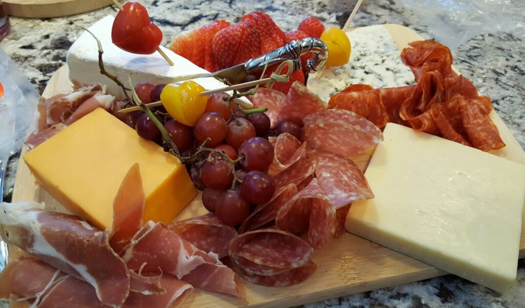 Alt text (115 characters): Wooden board with cheddar, white cheese, salami, prosciutto, ripe grapes, cherry tomatoes, and sliced strawberries.