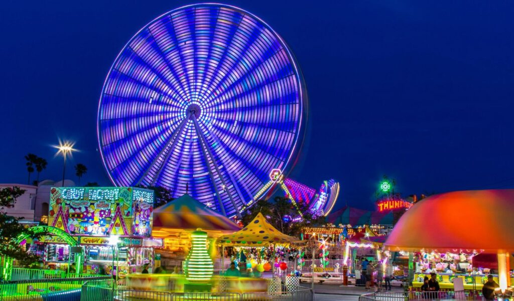 **Alt text:**
Colorful amusement park at night with a giant illuminated Ferris wheel, spinning rides, candy stalls, and vibrant neon lights creating a festive, lively atmosphere.