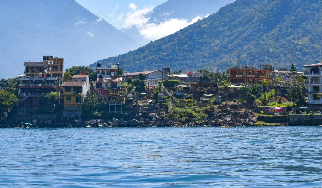 Colorful buildings line the shore of Lake Atitlán in Guatemala, backed by lush green mountains under a clear blue sky.