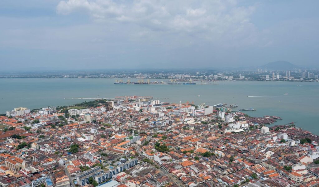 Aerial view of Georgetown, Guyana, featuring densely packed buildings with red rooftops along the coastline and a port area with docked ships under a cloudy sky.