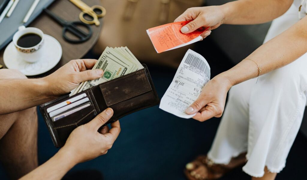 A person holding a wallet with cash and cards prepares to pay another person who is holding receipts, with a cup of coffee and scissors visible on the table in the background.