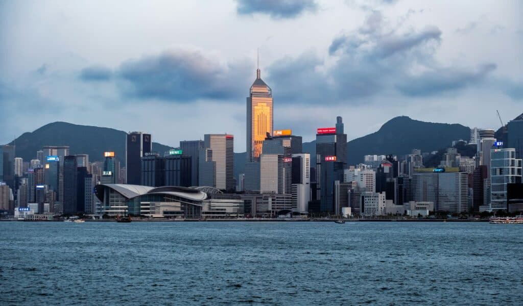 Scenic view of the Hong Kong skyline at dusk, featuring modern skyscrapers, Victoria Harbour in the foreground, and surrounding mountains under a cloudy sky.
