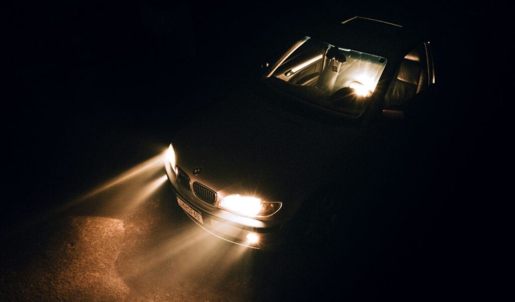 A car with its headlights turned on is seen in the dark from an elevated angle, casting bright beams of light onto the road ahead. The interior is dimly lit, highlighting the seats and dashboard.