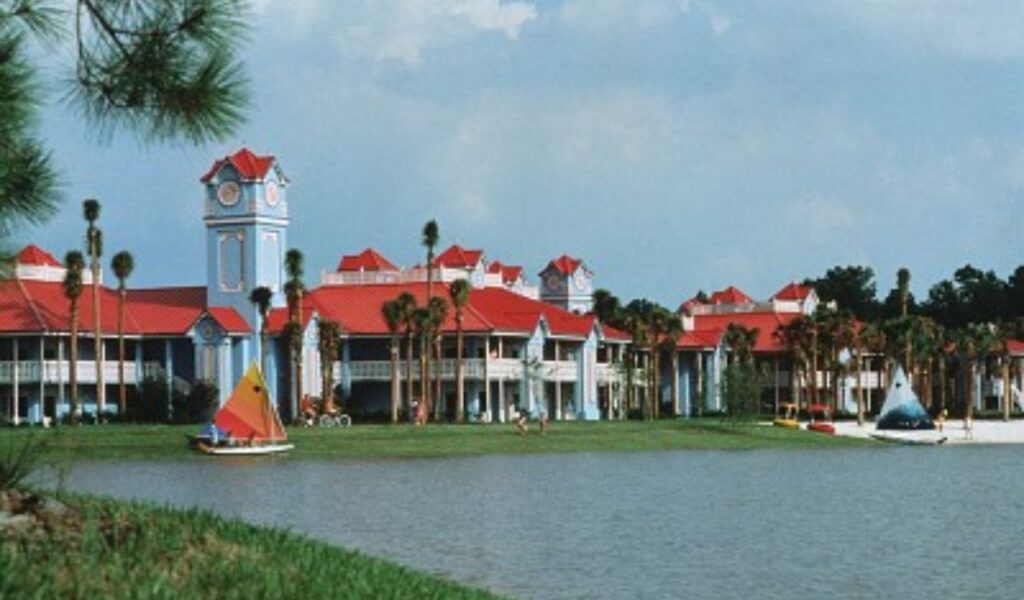 "View of a tropical resort with colorful buildings, palm trees, and a scenic lake, featuring outdoor activities like sailing.