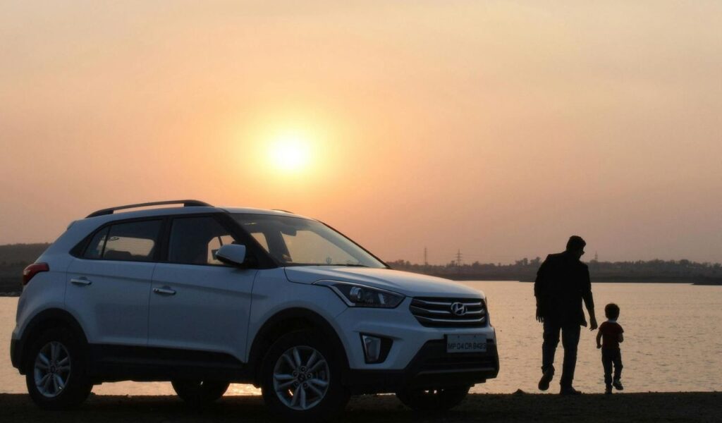 A man and child walk by a parked white SUV near a lake at sunset, with the glowing sun casting a warm light over the scene.
