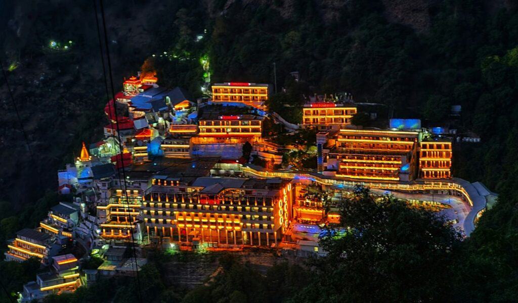 Vaishno Devi shrine complex brightly illuminated at night, nestled in the forested hills of Jammu, with buildings glowing in warm orange and yellow lights.
