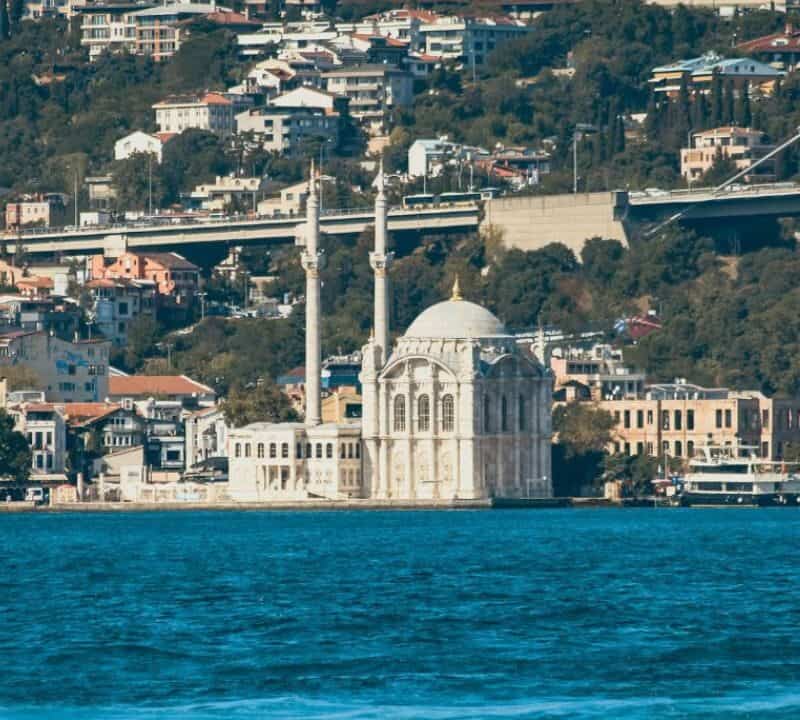 A mosque with two tall minarets sits along the waterfront in Istanbul, Turkey, with hillside homes and a bridge in the background.
