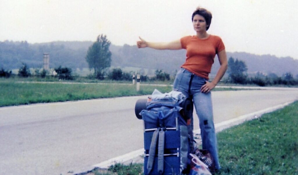 Young woman hitchhiking on a rural road with a large backpack, reflecting 1970s solo travel culture and perceived safety.
