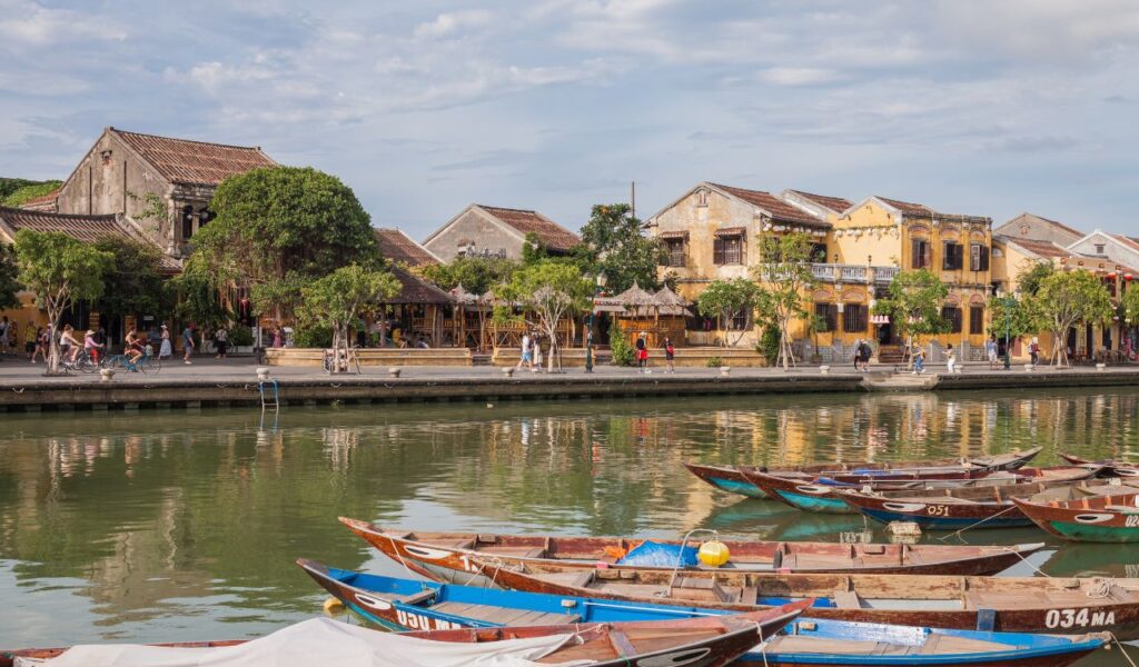 Traditional wooden boats on the Thu Bon River in Hoi An, Vietnam, with historic yellow buildings and lantern-lit streets in the background.